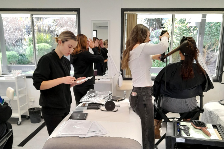 Three hair and beauty students work with a client, styling her hair and make-up. The client is seated in a chair with a black cape to protect her clothes.
