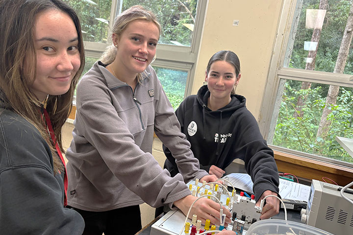 Three female electrotechnology students, Sarah Burke, Tayla Jones, Lily Bolton stand close together in a classroom setting. They hold wires attached to specialised equipment.