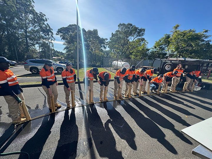 A group of students in high-visibility orange and navy workwear stand in a line outdoors, holding long-handled tools over a wet surface during a practical training session.