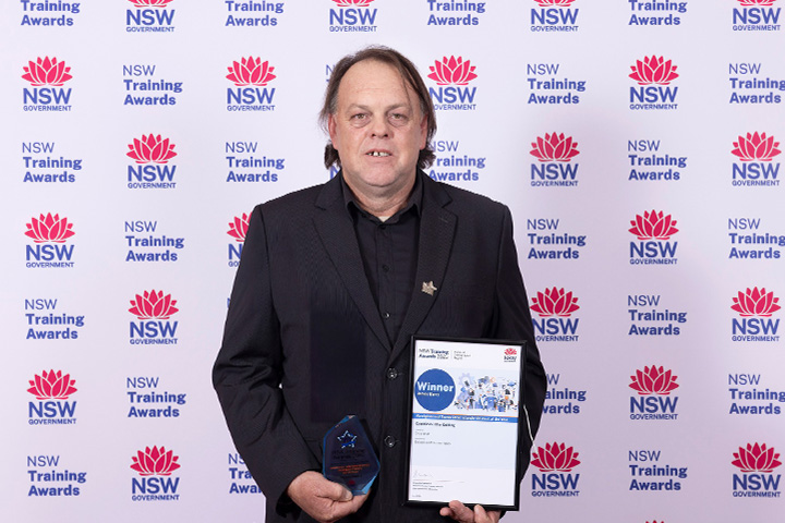 TAFE NSW graduate James Barry wears a black suit and shirt and stands in front of a NSW Government Training Awards backdrop holding a glass award and framed ‘Winner’ certificate.