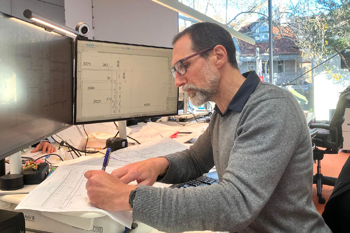 Jan Ellis, dressed in a grey jumper with a black collar and glasses, sits drafting at a busy desk in front of two large monitors displaying technical information. 