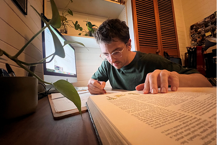 A man wearing glasses and a green tshirt writes in his workbook. He sits inside his bedroom.