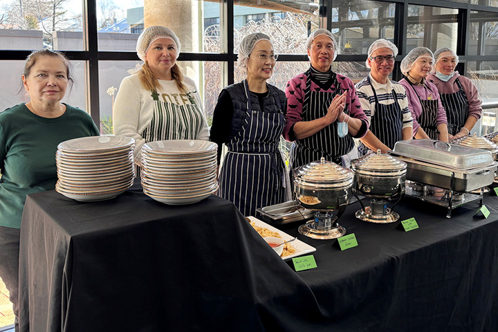 Seven TAFE NSW students stand behind a counter that has two stacks of plates, soup pots and other food serving utensils. Six of them are wearing hair nets and aprons. Five of them are looking at the camera and two are simply looking elsewhere. 