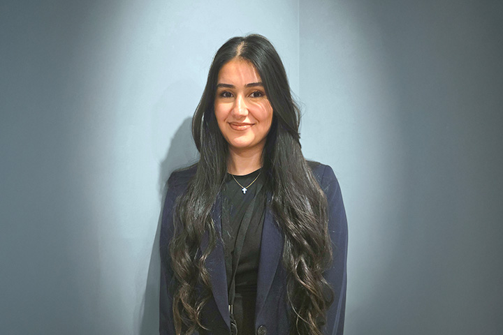 Rita Al-Ton with long wavy hair wearing a dark blazer over a black top and a necklace with a small pendant, standing against a plain background