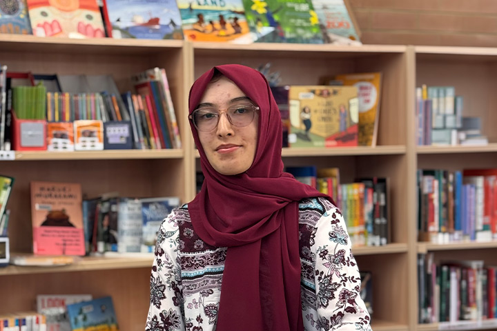 A young woman wearing a red hijab, a patterned blouse and glasses stands in a library with book shelves behind her.