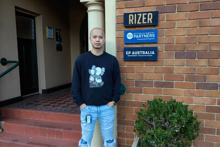 Themy Pham wearing a black graphic jumper and blue jeans, standing out the front of his work building. Beside him are signs that read “RIZER”, “Entertainment Partners” and “EP Australia”. 