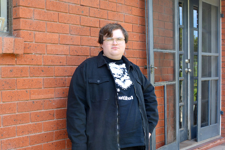 Smiling TAFE NSW Bega graduate, Thomas Rixon, stands outside dressed in black jeans, t-shirt and jacket, ahead of starting his university studies.