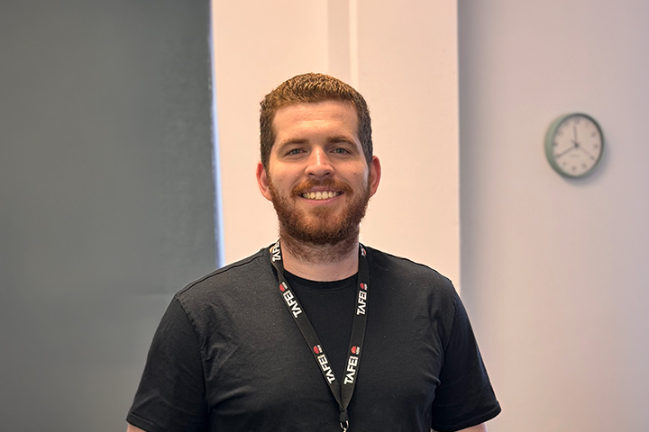 Tom Doe, with short light brown hair and a facial beard, stands smiling in a classroom setting. He wears a black TAFE NSW lanyard, with tables and chairs behind him. 