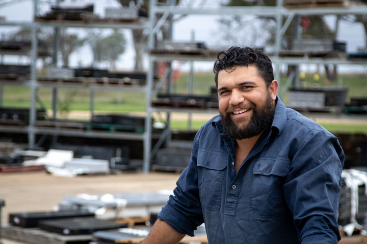 Travis Stahlhut, with short curly dark hair and a full beard smiles warmly while standing outdoors. He’s wearing a navy blue work shirt, with metal shelves and materials visible in the background, suggesting a trade or industrial setting.
