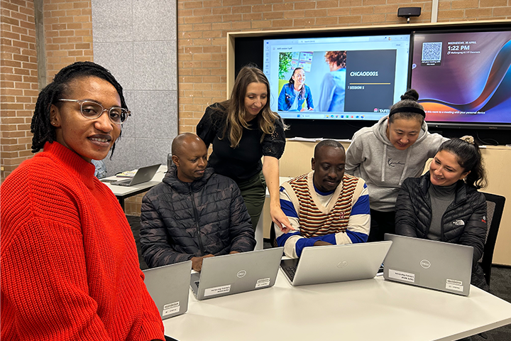 TAFE NSW teacher, Zeljka Jovanovic, wearing a bright red knit jumper, working with a group of migrant students who are all looking at their laptops