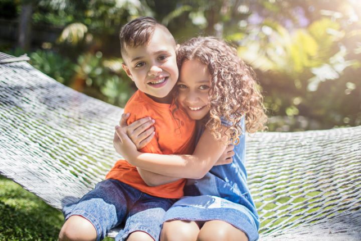 Two small children of Aboriginal heritage hugging each other while smiling. The boy has neat, short brown hair and wears an orange t-shirt and denim shirts, while the little girl with curly hair wears a light blue denim dress.