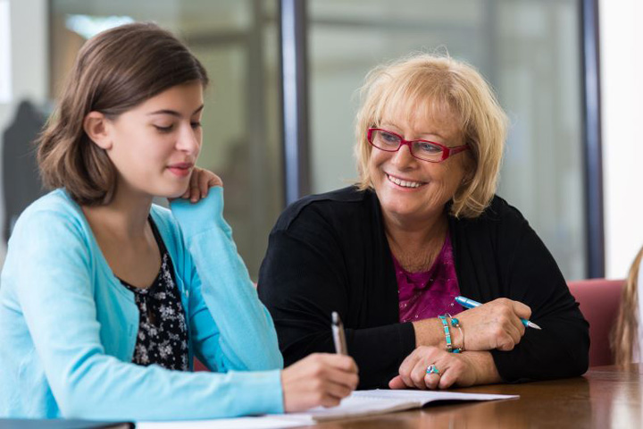 A teacher with short blond hair and glasses is sitting smiling beside the student next to her. The student is holding a pen and writing in her workbook.