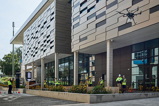 A group of workers dressed in hi-vis jackets stand outside the ground floor of a building testing drones, with one drone actively flying in the air.