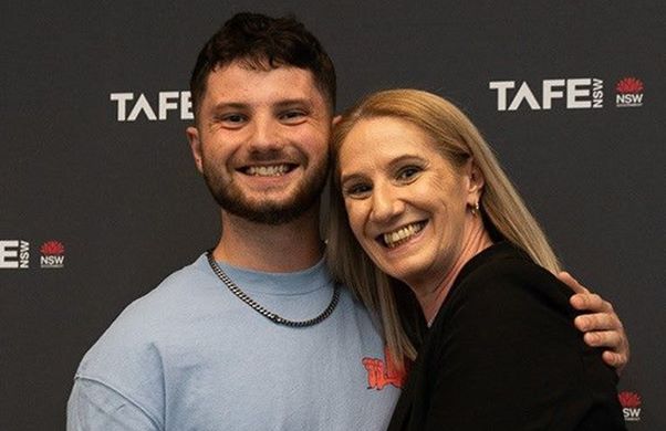 Caleb Sturges, a 20 year-old nursing graduate at TAFE NSW Kingswood, smiles happily. There is a TAFE NSW banner board in the background. 