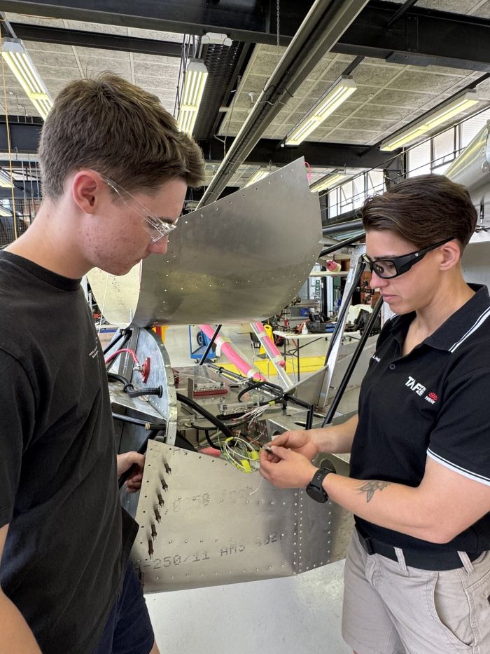A TAFE NSW instructor in safety glasses and a black TAFE NSW shirt is working alongside a student on the wiring of an aircraft component. They are standing in an aviation workshop with tools, equipment, and parts of an aircraft visible in the background.