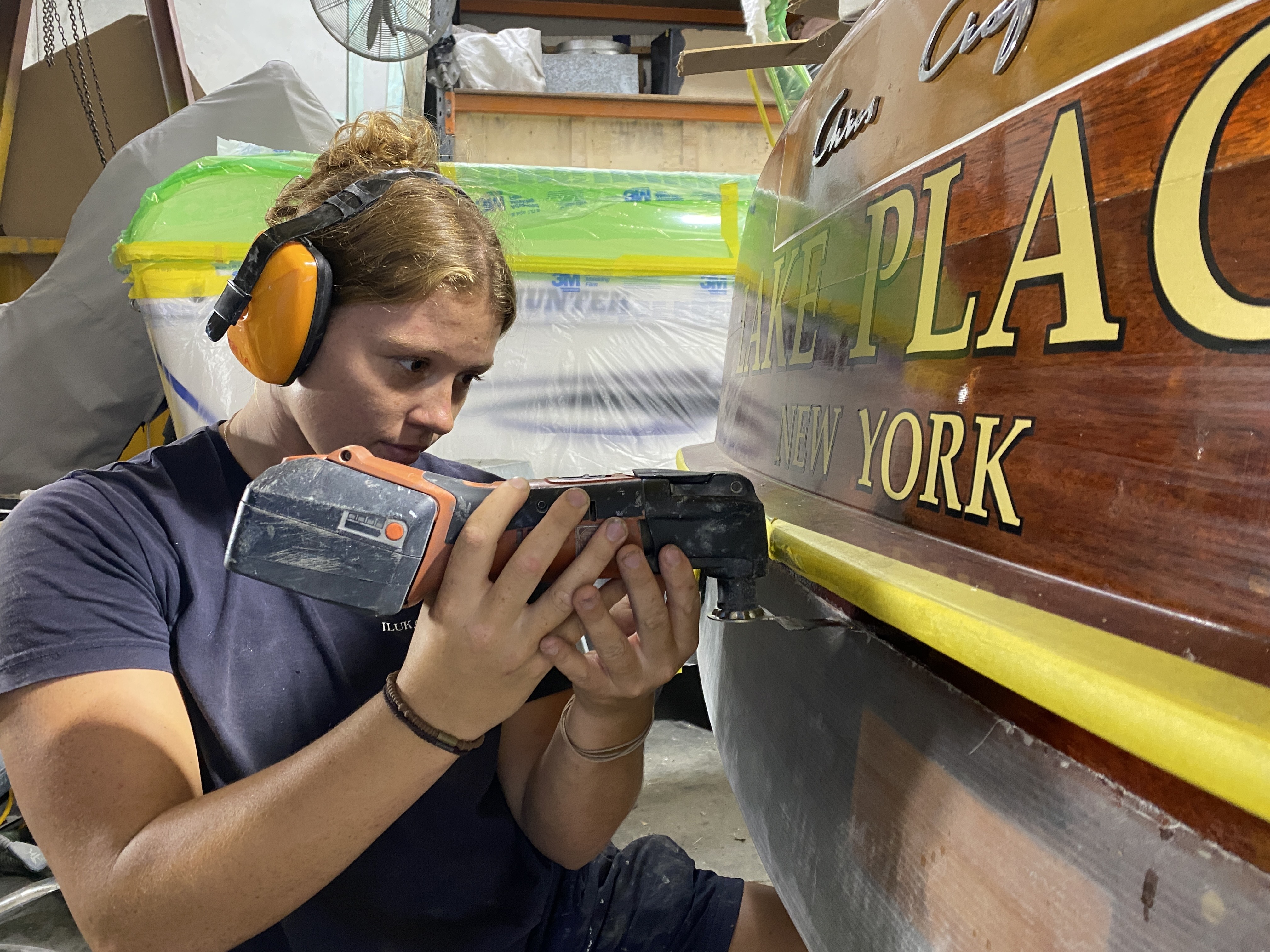 Talia Dunning working on the construction of a boat. She has earmuffs on and a power tool in her hands. 