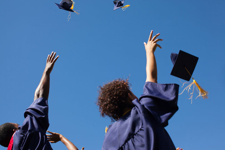 Two graduates, dressed in dark blue gowns, throw their yellow tasseled caps into the air in celebration. Their arms are outstretched against a clear blue sky.
