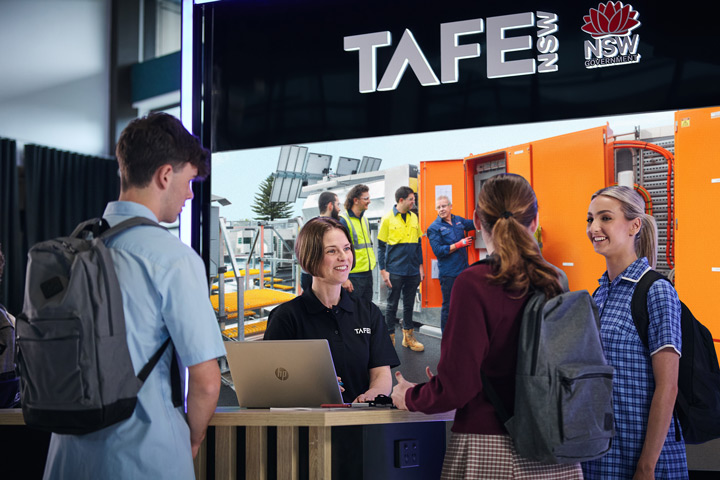 TAFE NSW staff member engages with students at an expo stand featuring a large screen displaying electrotechnology training, including solar panels and industrial equipment, with NSW Government branding visible.