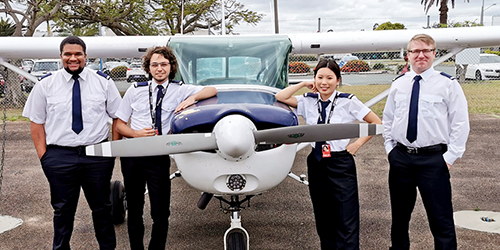 Four aviation students wearing pilot uniforms stand in front of a single-engine training aircraft, representing flight training and pilot education.