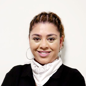 Farhana Dawood is smiling at the camera against a white wall and she is wearing large hoop earrings, a white scarf and a black top.