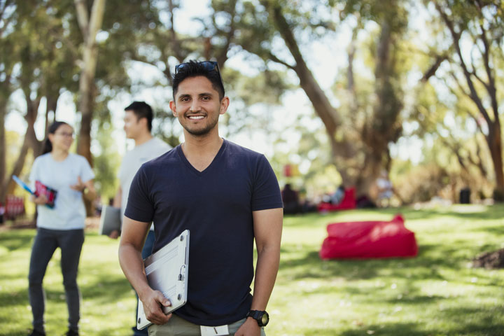 A young student is standing outside on a campus lawn, holding a laptop and smiling at the camera. There are more students in the background talking while they are standing or lying under some huge trees.
