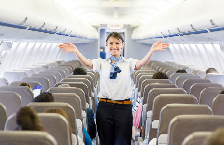A female flight attendant is standing in the aisle of an airplane wearing a uniform and indicating where the exists are with her arms. There are many passengers seated around her.