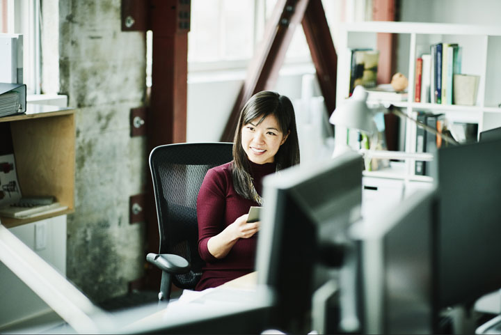 A professional woman in a maroon sweater sits at a desk in an office, smiling while holding her phone.