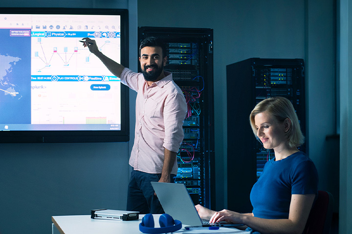 Two professionals in a server room analyzing data on a screen with charts and graphs.