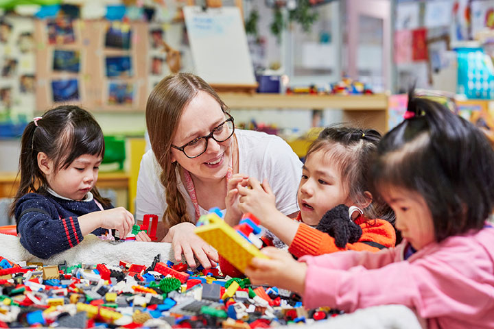 Young children engaged in an early childhood education activity with colorful building blocks, guided by an adult in a classroom setting.