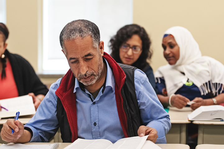 Adult students in a classroom setting, engaged in writing or reading with textbooks open in front of them.
