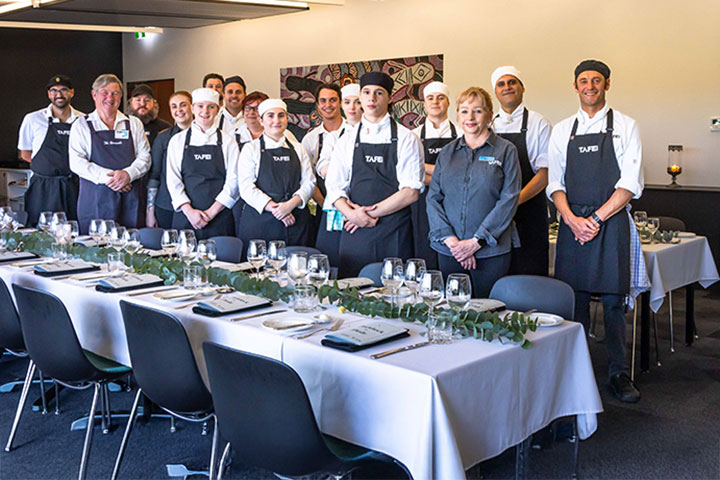 A group of chefs and students from TAFE NSW, dressed in black aprons and chef uniforms, stand proudly behind a beautifully set dining table. The table is adorned with elegant tableware and a eucalyptus leaf arrangement, ready for a formal dining event. The group is posing in a well-lit, modern room, with a background featuring artwork and minimal decor.