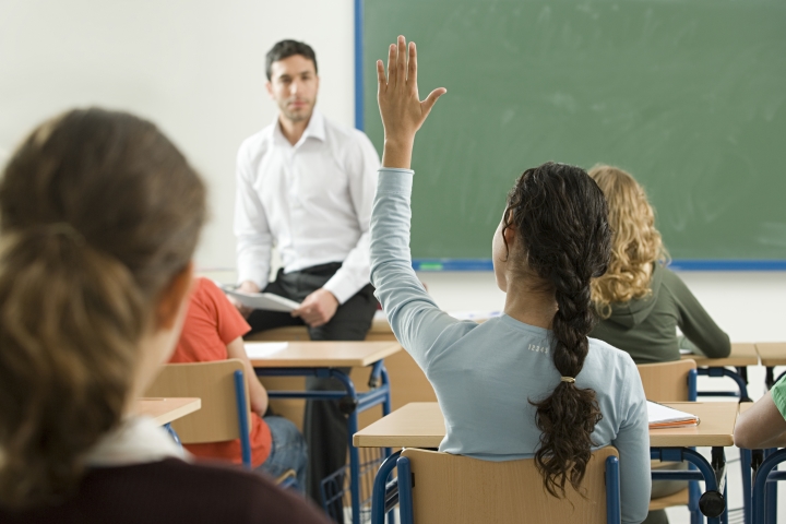 A group of students sit in a classroom with the teacher sitting on the edge of his desk at the front of the class. One student has her hand raised.