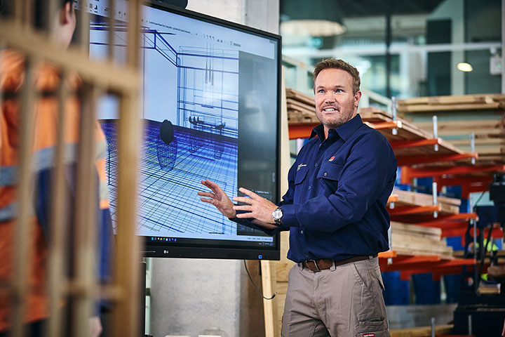 A construction teacher at the Institute of Applied Technology stands in front of a large screen displaying a 3D construction model, explaining its features in a classroom with shelves of building materials.