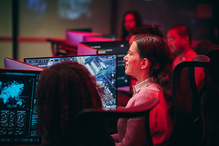 A group of professionals are seated at computer workstations in a control room, monitoring large screens displaying maps and data visualisations
