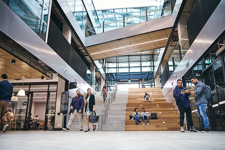 Modern, light filled campus atrium with people in transit, standing in conversation and some seated on the bench side of a wide stairway.