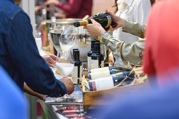 A wine market tasting scene with wine bottles decoratively displayed and one person pouring a glass of wine for another.