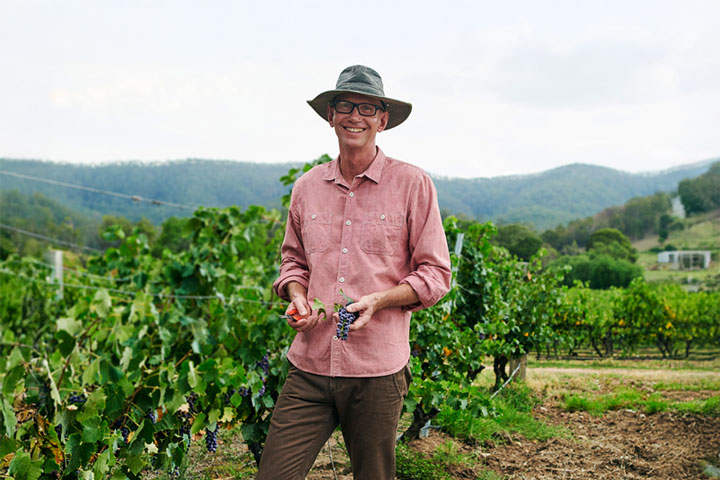 A man stands smiling proudly at the camera, holding a bunch of grapes. He’s surrounded by a lush and healthy vineyard. He’s wearing a bright collared shirt, long, brown pants and boots, and a wide-brimmed sun hat.