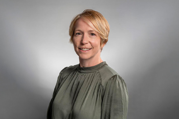 Helena Edgerton, with short, blonde hair wearing a long-sleeved, dark green blouse. The background is a plain grey gradient. 