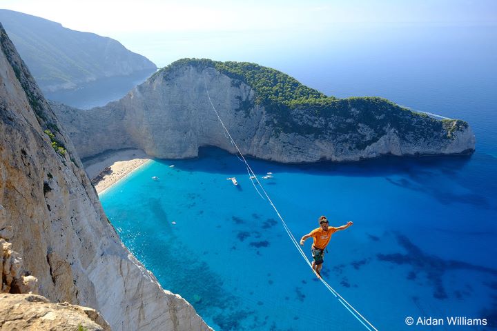 A high-angle photograph by Aiden Williams shows a person in an orange top and shorts tightrope walking between two mountains. In the background, a scenic beach cove features white cliffs adorned with greenery.