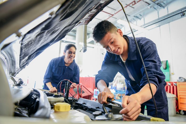 Two young mechanics in blue coveralls work together under the hood of a car in an auto repair setting. They are smiling and are engaged in the repair process.