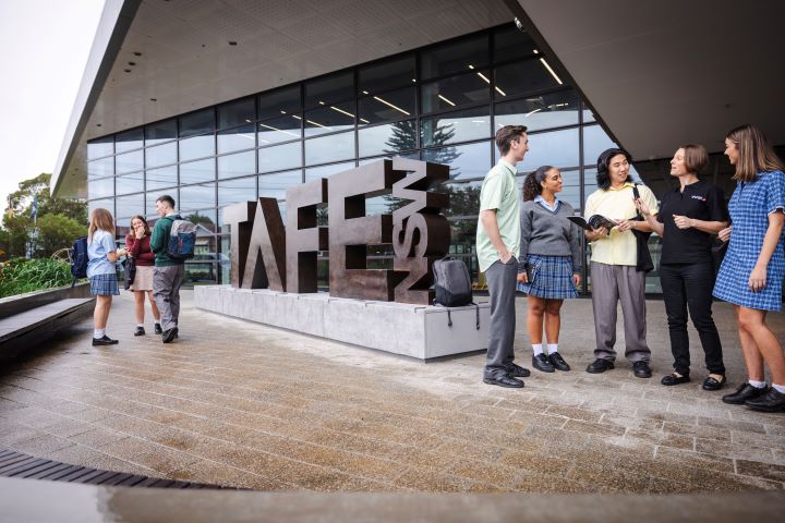 A group of school students stand outside a modern building with a large 'TAFE NSW' branded sculptural signage.