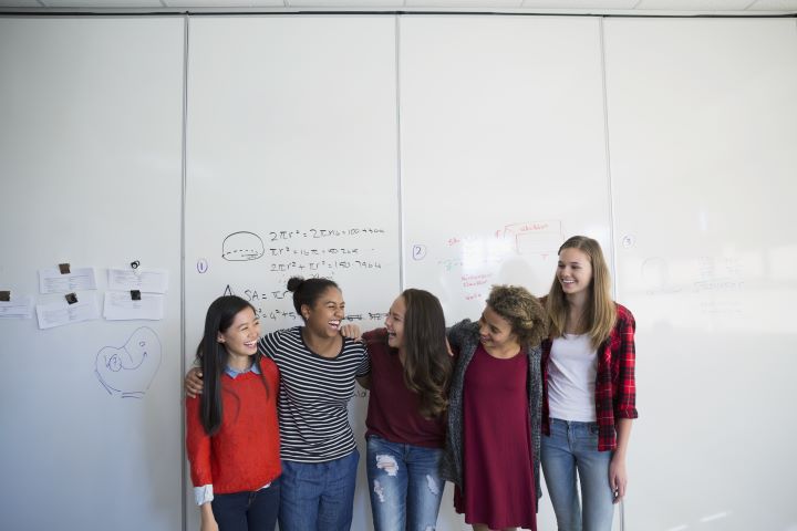 A group of women standing together, smiling and admiring their achievements. They are standing in front of a whiteboard with mathematic equations. 